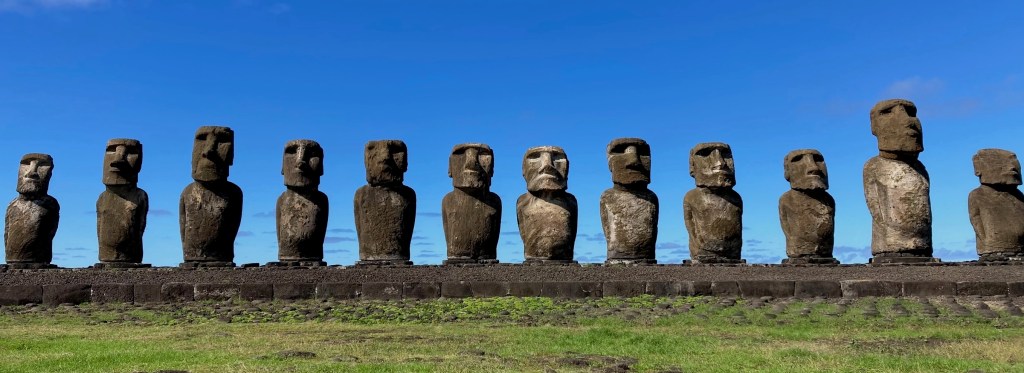 photo of twelve grey stone statues with large heads and torsos with blue sky behind and green grass in front