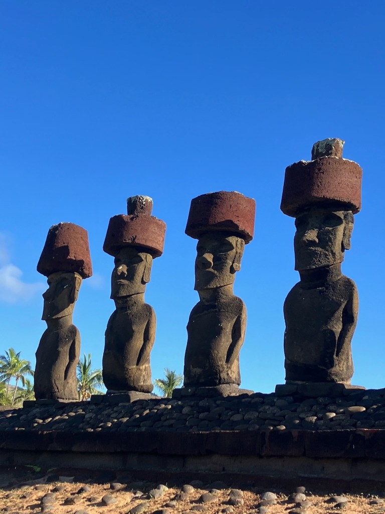 photo of four gray stone statues with large heads and torsos and large red stone "top knot" caps with blue sky behind them
