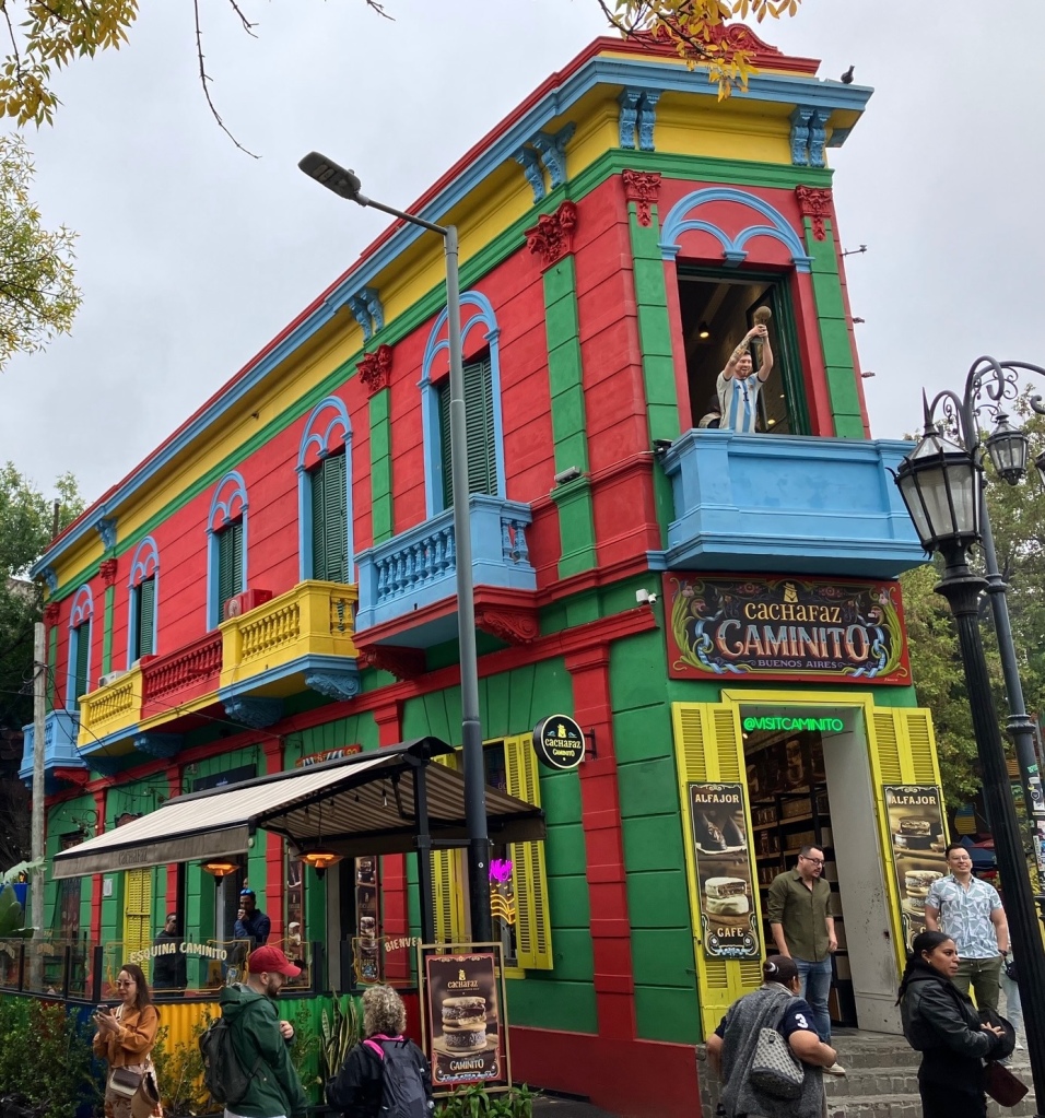 photo of a red, green, yellow, and blue two-story building, only one room wide at the front. In the front upper balcony stands a statue of Lionel Messi wearing his blue-and-white Argentina jersey and holding the World Cup soccer trophy over his head.