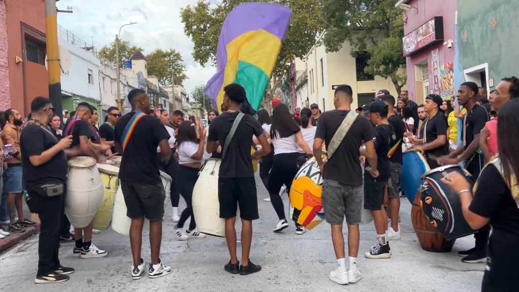photo of a group of young men wearing black t-shirts with large bongo-shaped drums strapped over their shoulders. They are standing in a circle with their backs to the camera, while a circle of young women in white t-shirts dances in the middle and a purple and yellow flag waves behind them.