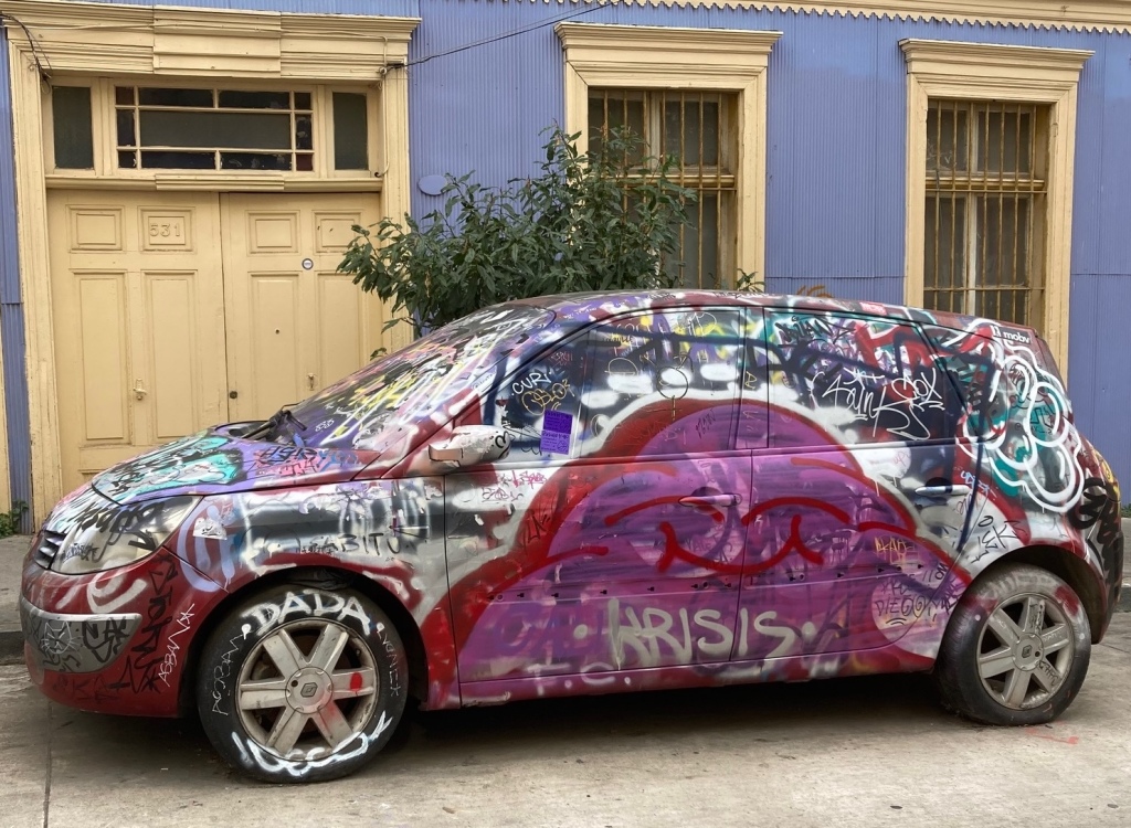 photo of a small car covered in silver, red, purple, and black spray-painted graffiti and parked in front of a purple house with yellow doors and window frames