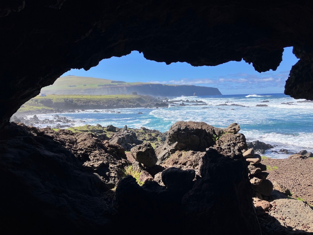 photo looking out of a dark craggy cave with gray rocks in the foreground and blue-green sea with white waves and green-topped cliffs in the background
