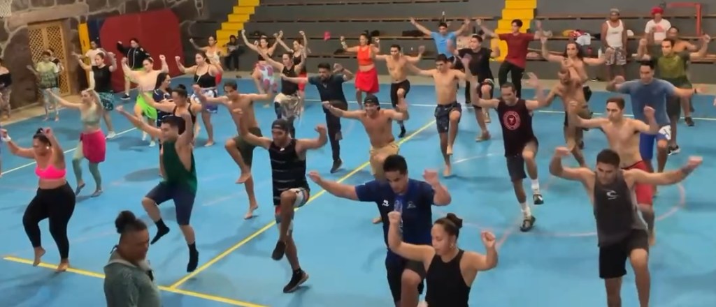 photo of dozens of people dancing in unison, arms and legs raised, wearing shorts and t-shirts on a blue gym floor.