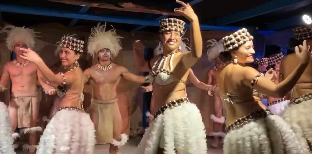 photo of four Polynesian women wearing white feather dresses, waving curved arms and hands and smiling broadly. Behind them are two Polynesian men wearing loin cloths and feather headdresses, dancing with arms outstretched.