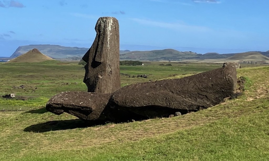 photo of one large gray stone head standing up behind another stone head and torso lying face down in the green grass
