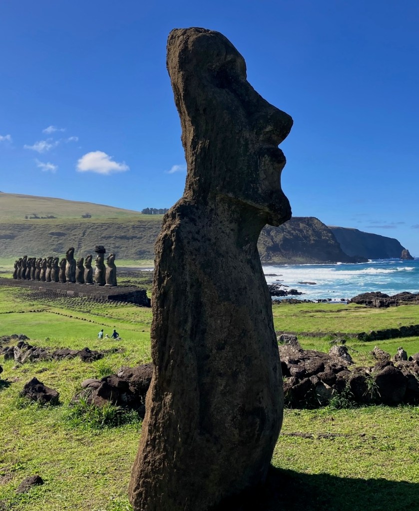 photo of a large gray stone head and torso standing on green grass. In the background are 15 similar statues near a beach with waves crashing.