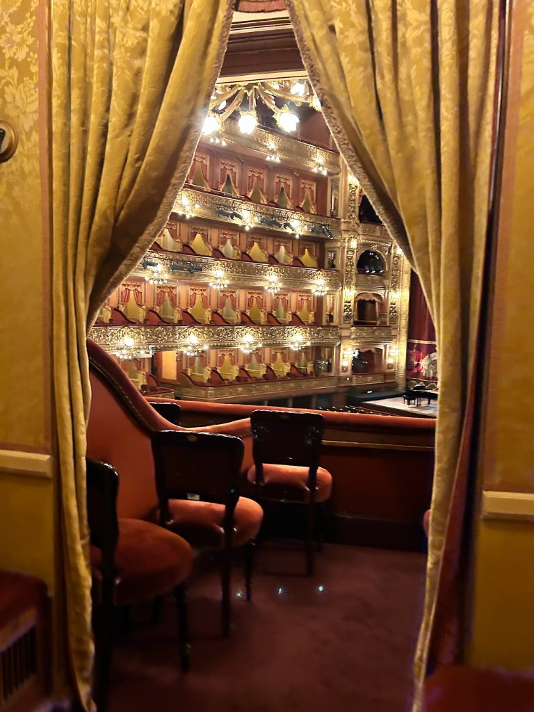 photo of golden curtains in a doorway, pulled back to reveal red-and-dark-wood chairs with a balcony in front of them and a view of illuminated gold balcony boxes.