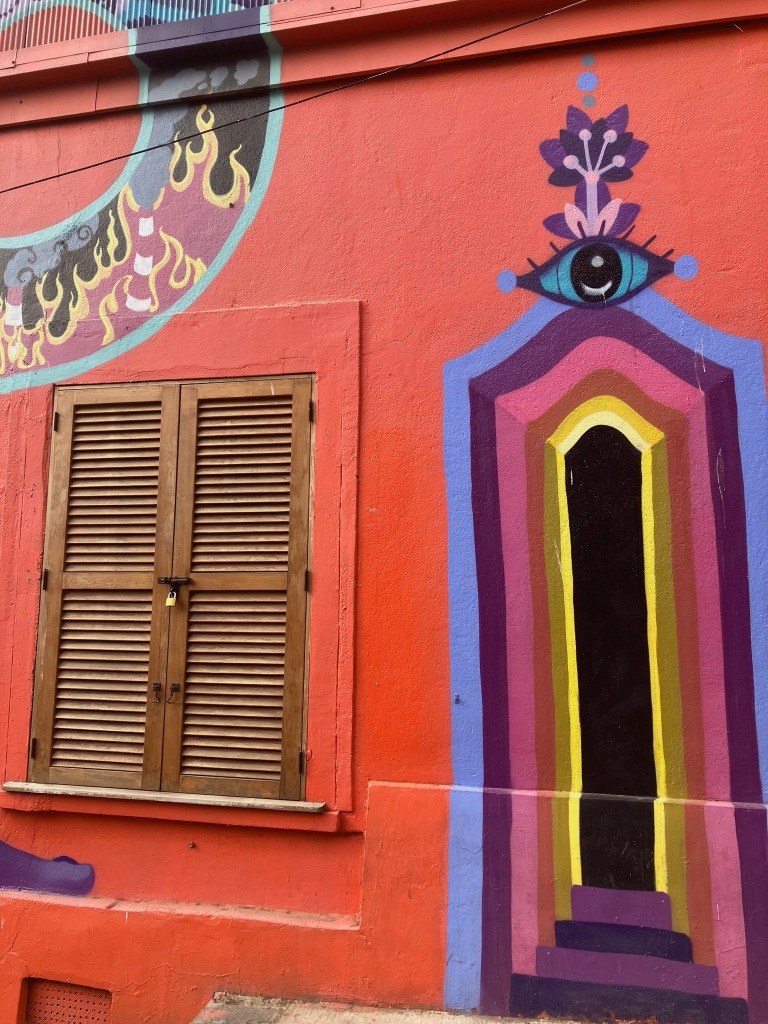photo of a red wall with a window with dark wooden shutters and a painting in blue, purple, red, and yellow that creates an illusion of an entry doorway