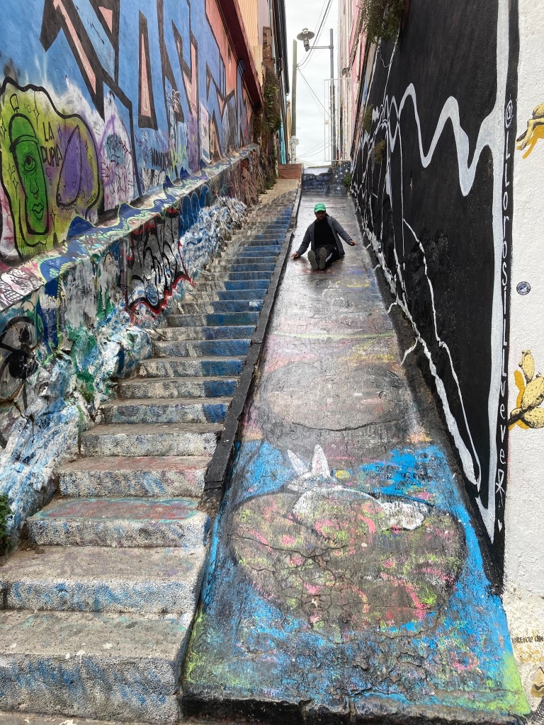 photo of a steep stone staircase next to an equally steep smooth stone surface. All surfaces are covered in colorful street art and the a person is sliding down the smooth area.