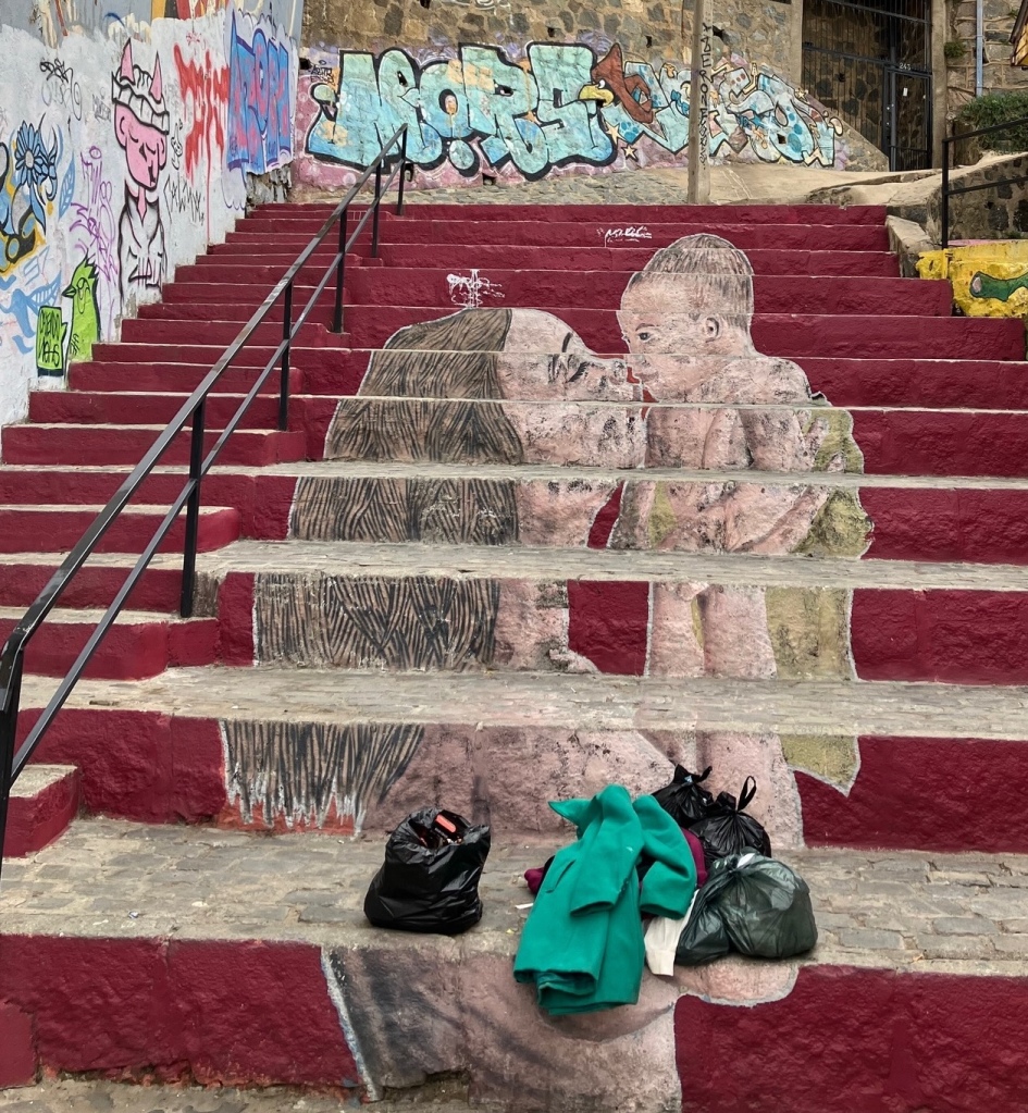 photo of a stone staircase with red risers and an image of a woman kissing a baby painted on the risers. Graffiti covers the walls and a pile of clothes and plastic bags sits on the bottom step.