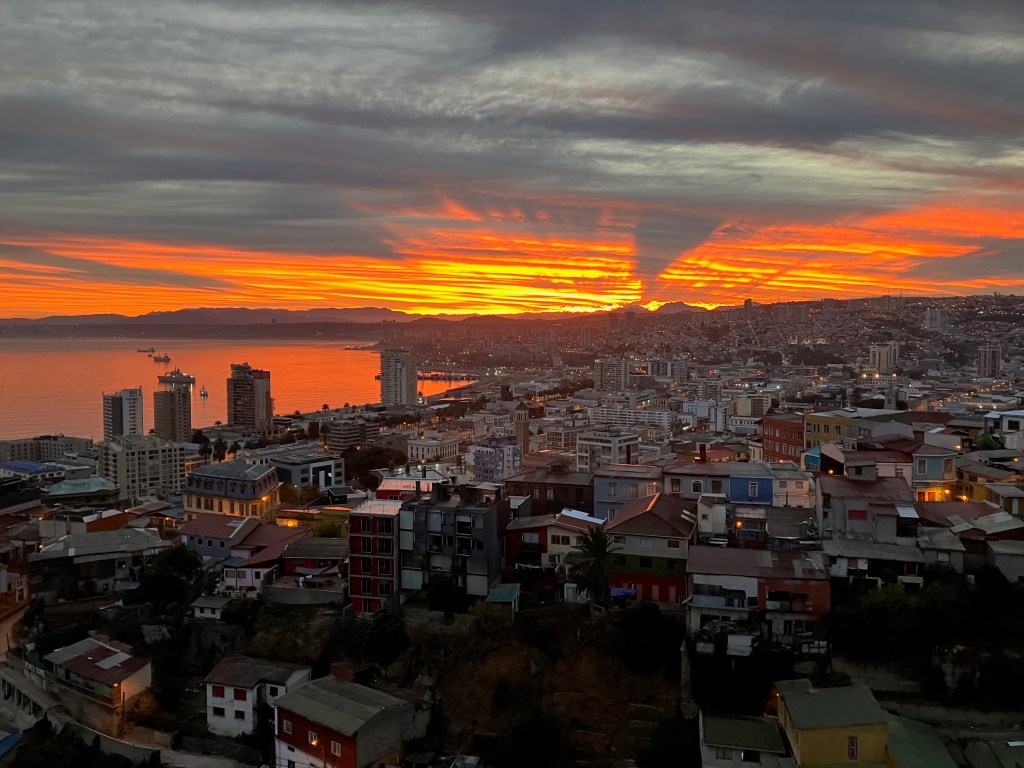 photo of orange and red streaked clouds in the distance, across city buildings and reflected in a curved harbor