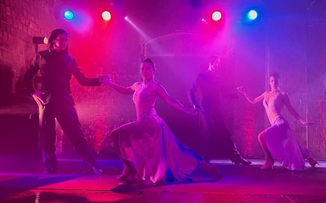 photo bathed in red and purple stage lighting of a man in a dark suit standing and holding the right hand of a woman wearing a white dress, with her left left leg crouched and her left hand and right leg extended back. Another dancing couple mirrors their position in the background.
