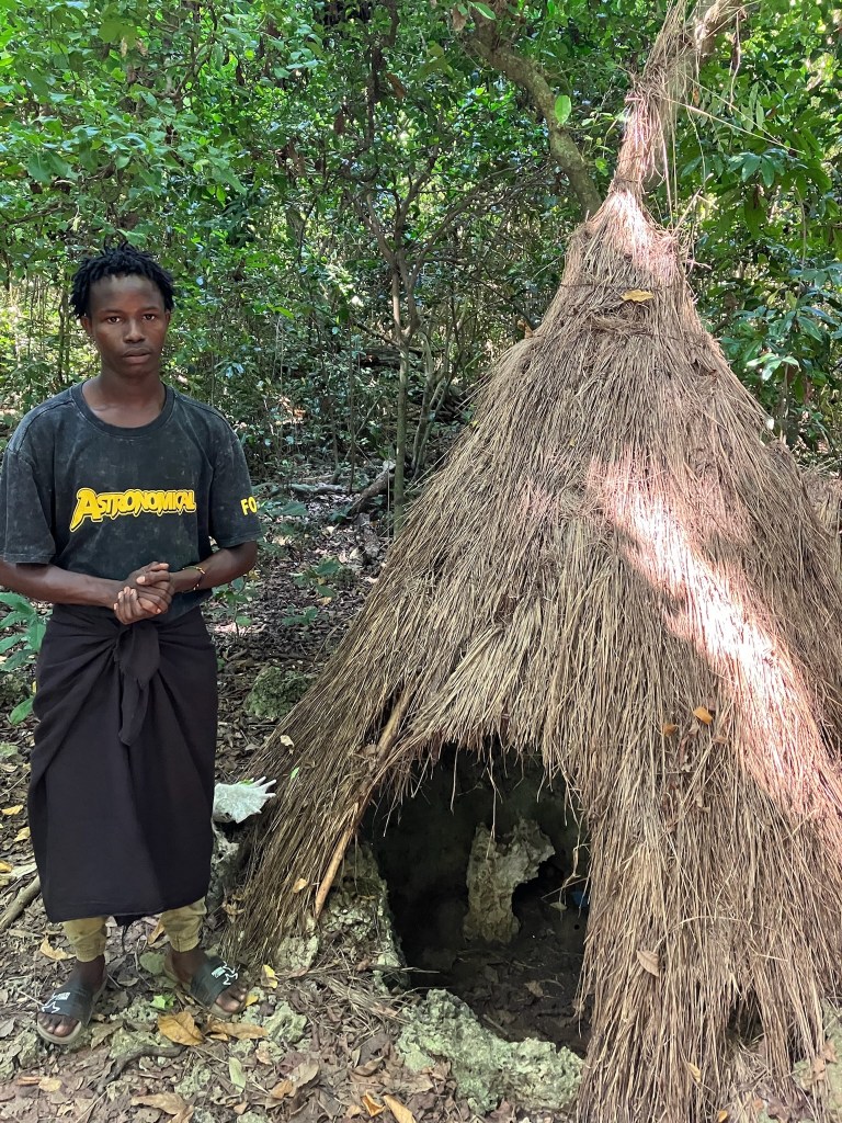 Photo of a young African man in a dark t-shirt and black skirt standing next to a thatched, conical hut with a small opening up to his hips