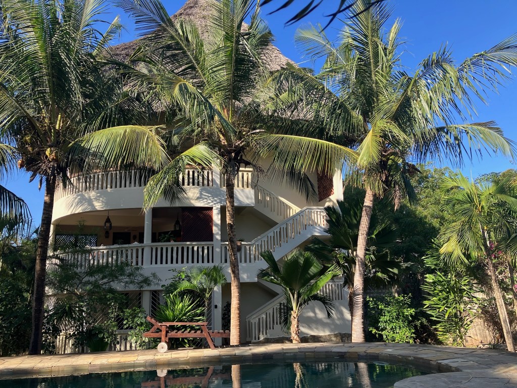 photo of a 3-story white building with balconies on each floor, a brown thatched roof, palm trees in front, and the edge of a swimming pool in the foreground