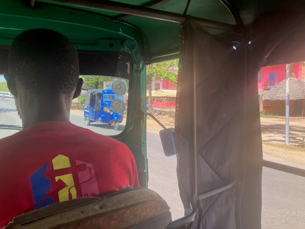 photo from inside a tuk-tuk of the back of the head of a man wearing a red t-shirt, driving a tuk-tuk, with a blue tuk-tuk visible through the windshielda