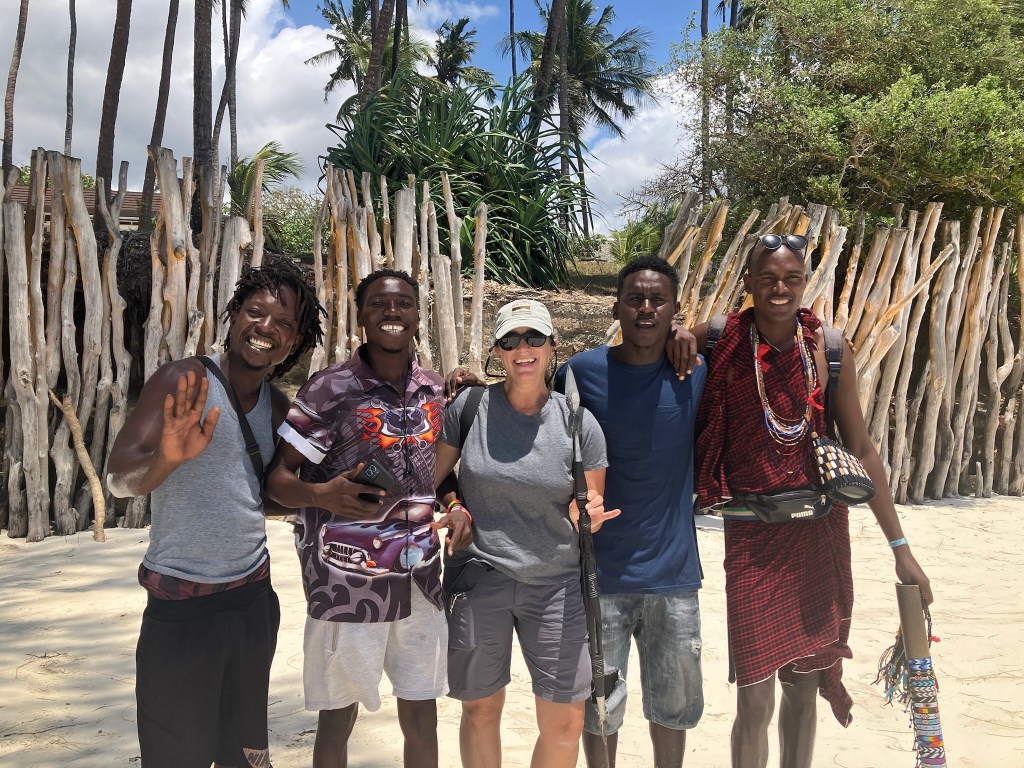 photo of four smiling African young men, one dressed as a Maasai warrier, with Rachel in the middle and a log enclosure in the background