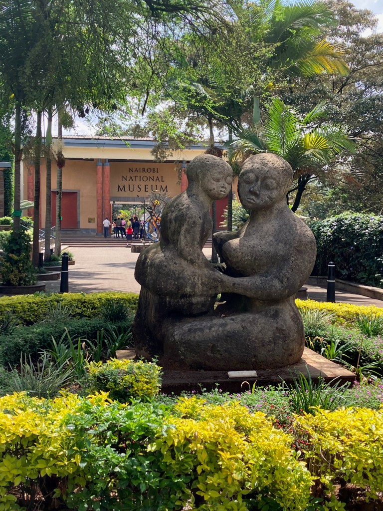 photo of the entrance to the Nairobi National Museum sign on a light brown wall, with a stone statue of a mother and child in the foreground