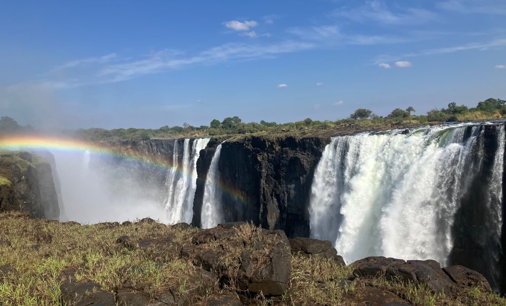 photo of a white waterfall spread along a dark cliff with a rainbow arching over it, green grass in the foreground, and blue sky in the background