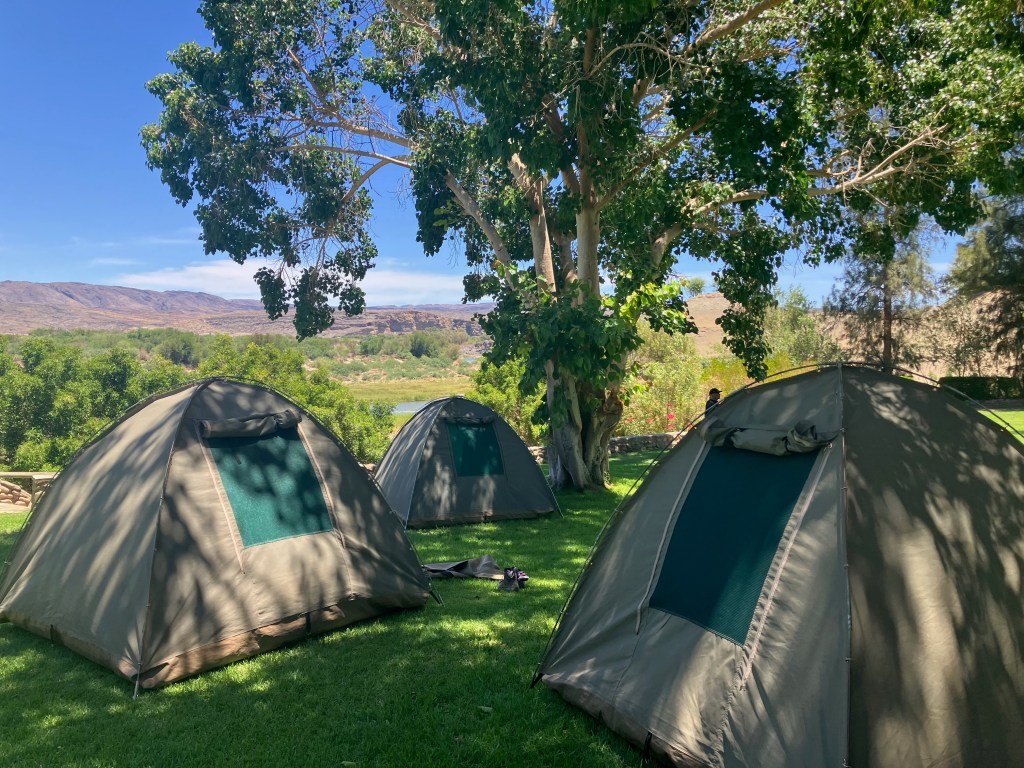 photo of 3 green dome tents on a green lawn with leafy trees overlooking a view of a river valley with brown mountains in the distance
