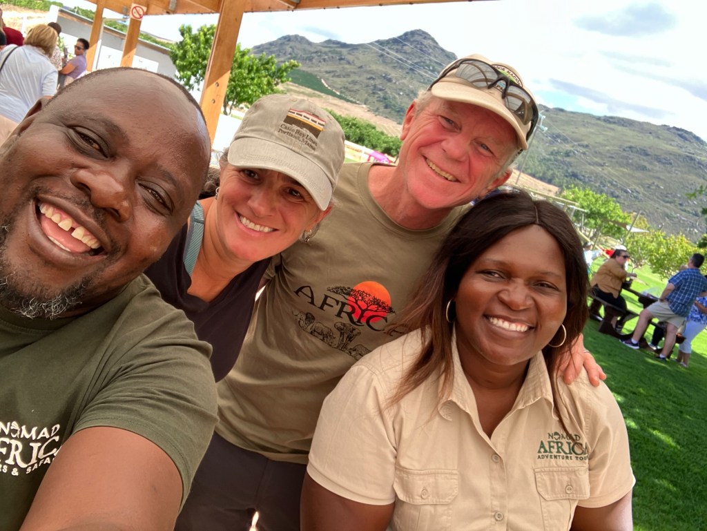 selfie photo of a smiling African man, a white woman and man, and an African woman, with mountains and people behind them