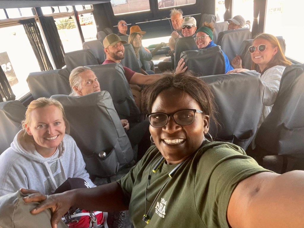 Selfie photo of an African woman wearing glasses, and in the background are bus seats occupied by 11 white people of different ages and genders