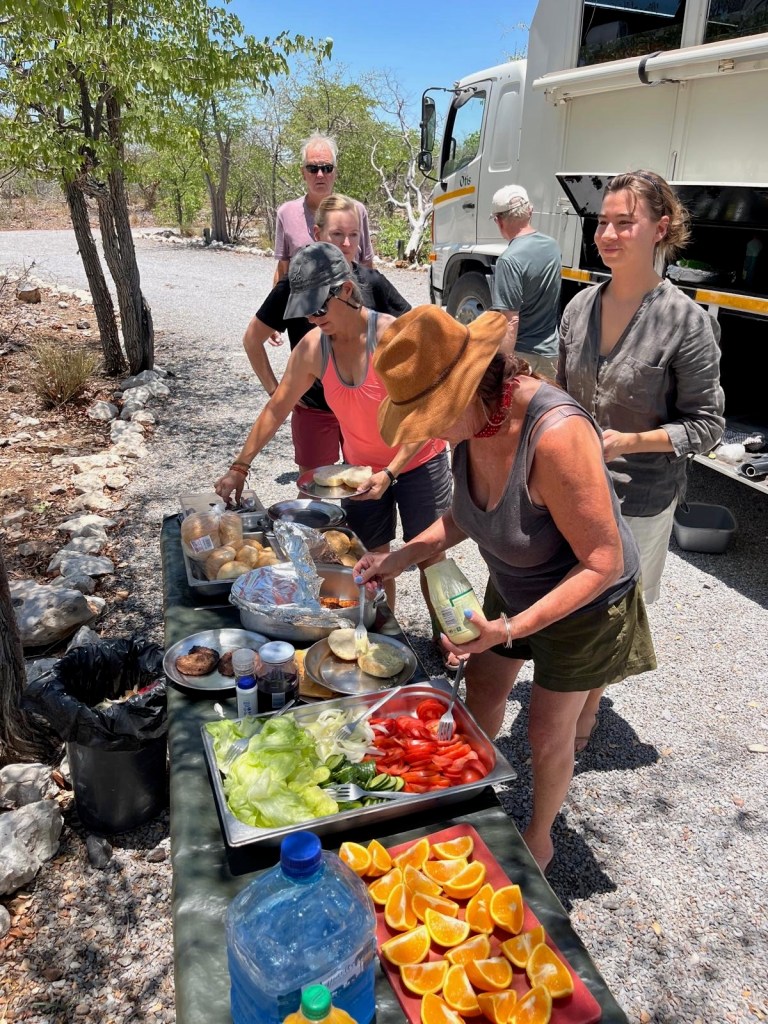 photo of 5 people lining up to put food on metal plates at a table with orange slices, red pepers, green lettuce, all next to a white truck with open storage compartments on the side