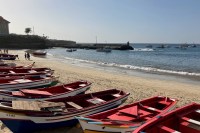 photo of several open-hulled wooden boats in red, blue, and white lined up on a sandy beach with small waves and a jetty behind