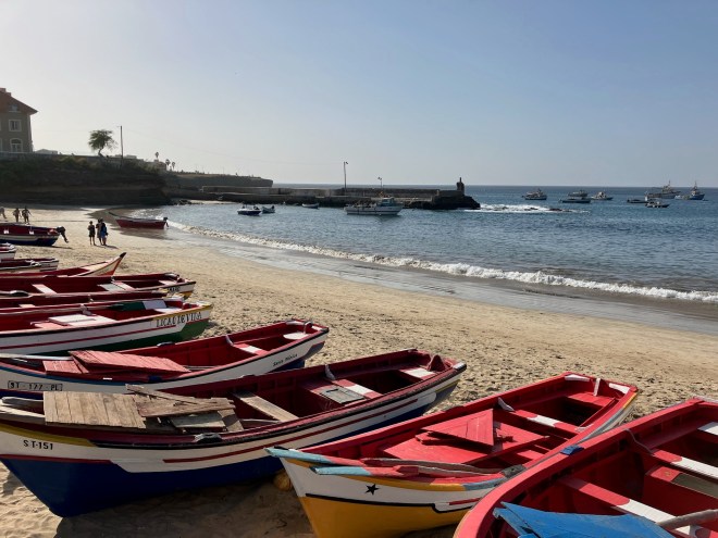 photo of several open-hulled wooden boats in red, blue, and white lined up on a sandy beach with small waves and a jetty behind