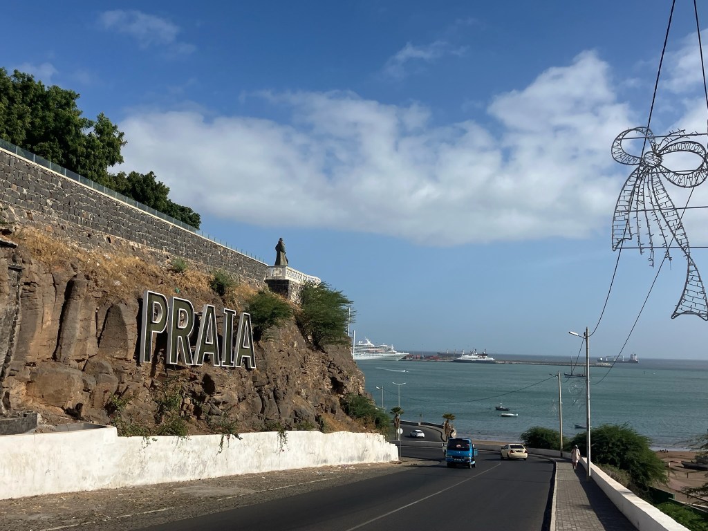 photo of a rock wall with PRAIA sign on it, a paved road in the foreground and blue-green ocen behind and a white cruise ship at a pier
