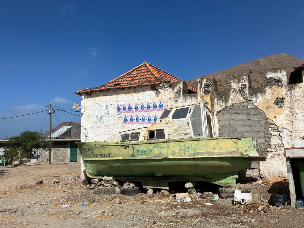 photo of a wrecked boat on blocks in front of a ramshackle cement house with a red tile roof