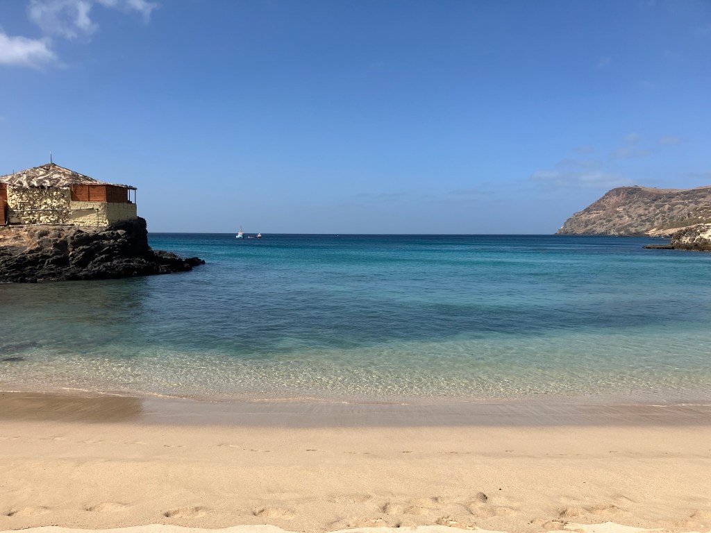 photo of a blue-green clear water cove with a rock shelter on top of rocks to the left and a mountain ridge to the right