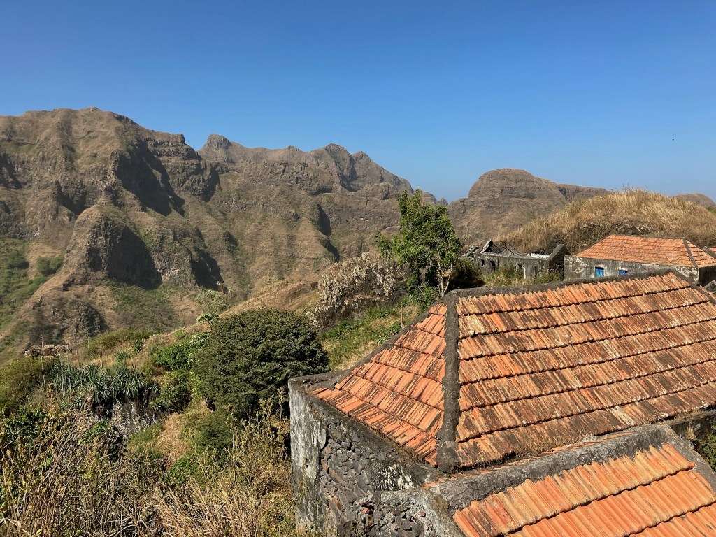 photo of a jagged mountain ridge with bright blue sky, looking across two dirty red-tile roofs