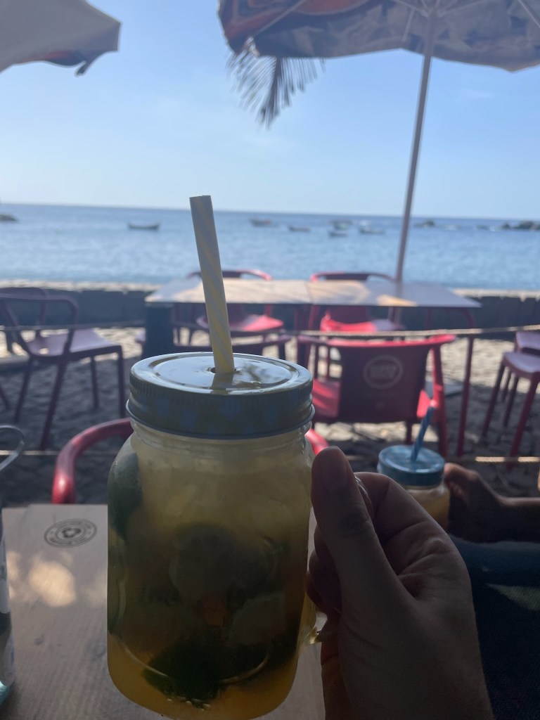 photo of a green drink in a jar with a straw on a table overlooking sand and a blue harbor