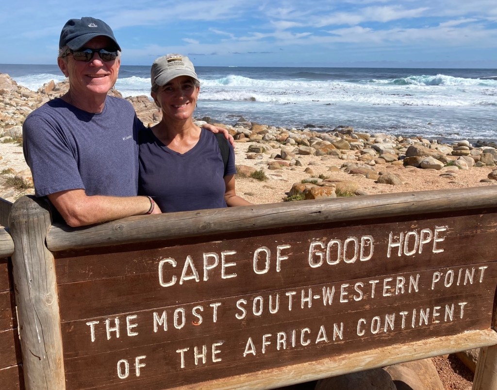 photo of Al and Rachel standing behind a sign that says Cape of Good Hope - The Most South-Western Point of the African Continent" with waves and a rock-and-sand beach behind
