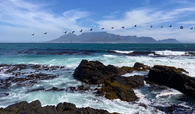 photo of a distant flat-topped mountain with buildings at the bottom, looking across green-blue ocean and white waves on jagged dark rocks, with a line of birds flying across