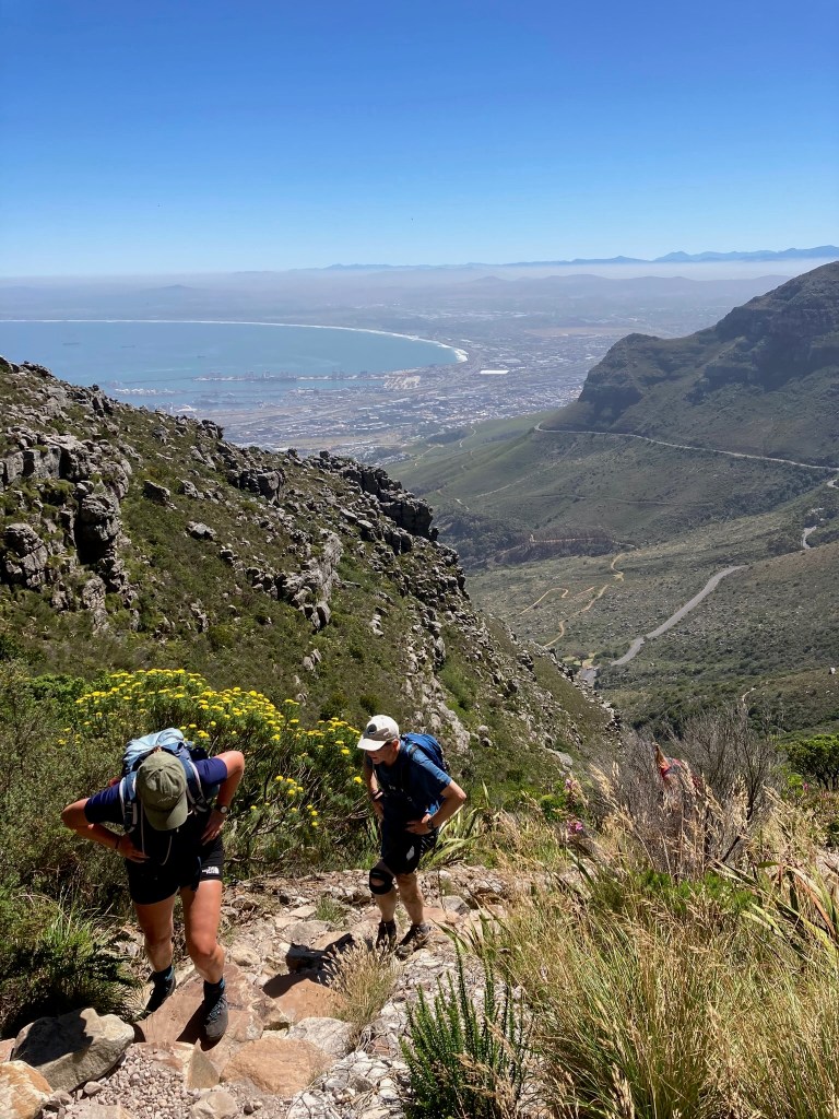 photo of a panoramic view across green and rocky mountains to a curved white beach in the distance with two people climbing up a steep trail toward the camera