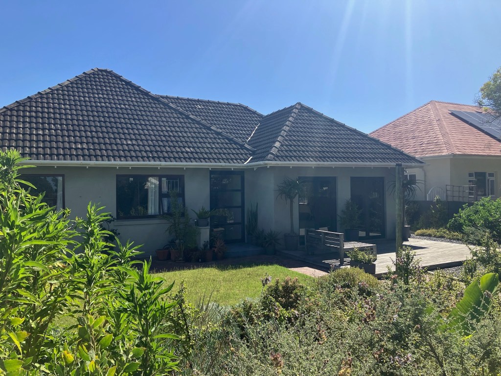 photo of a one-story house with white walls, large picture windows, a brown triangular tile roof and bushes in the foreground
