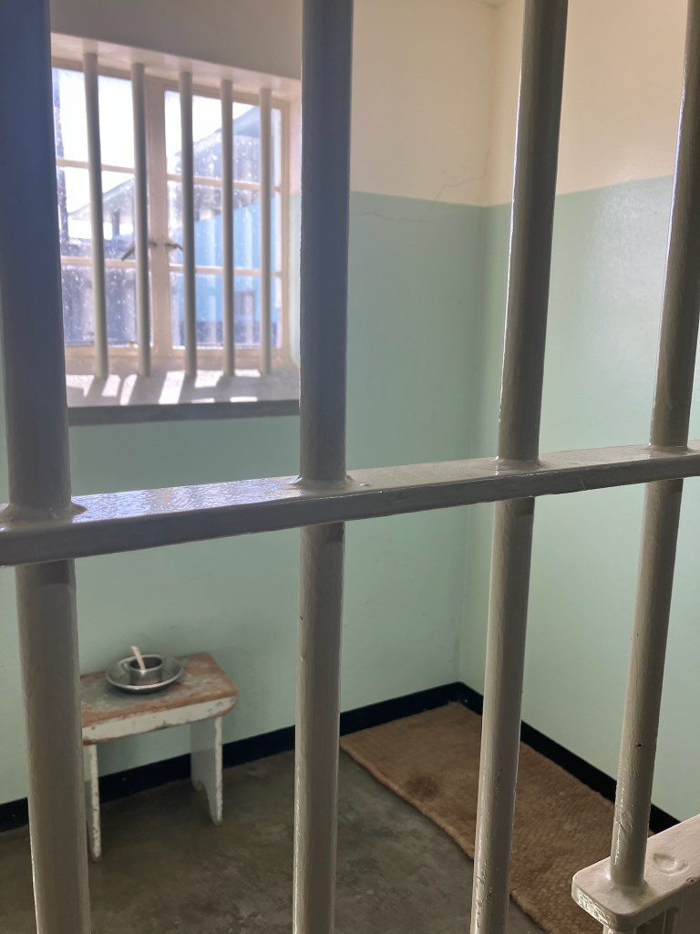 photo looking through the bars of a jail cell with one small table, a metal plate and cup, a brown mat on the floor, and a barred window behind