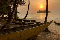 photo of the sun setting over a small tree-covered island, across the water from two narrow wooden boats resting on the beach under palm trees.