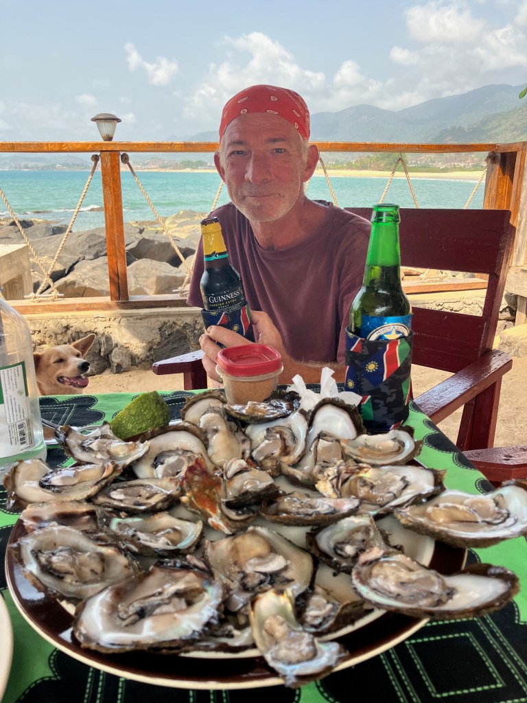 photo of a white man in a red bandana holding a beer bottle with a large plate holding 24 open oysters, with the blue-green sea behind hiim