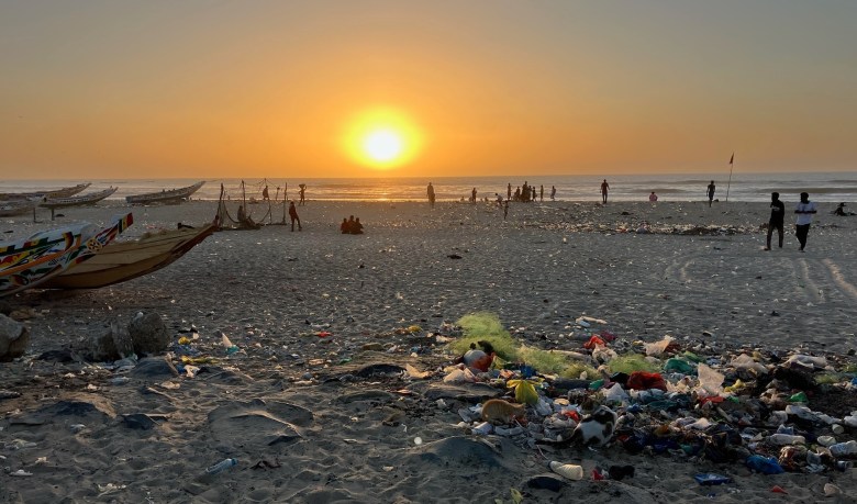 phot of the sun setting over the ocean, with silhouettes of people standing on the beach, wooden boats lined up on the left, and piles of trash in the foreground
