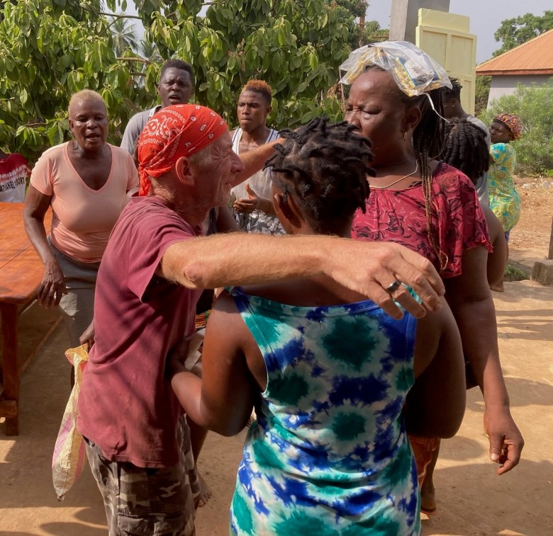 photo of a white man wearing a red bandana and maroon t-shirt hugging and dancing with two black women while 3 others in the background sing