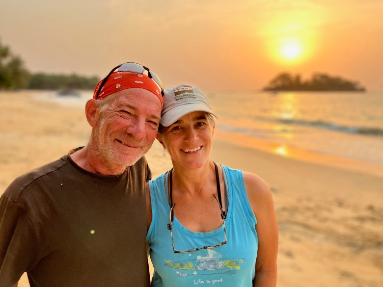 photo of a white man wearing a red bandana and black t-shirt and a white woman wearing a gray baseball cap and blue tank top, with a sandy beach, glittering water, a small tree-covered island, and the setting sun behind them.