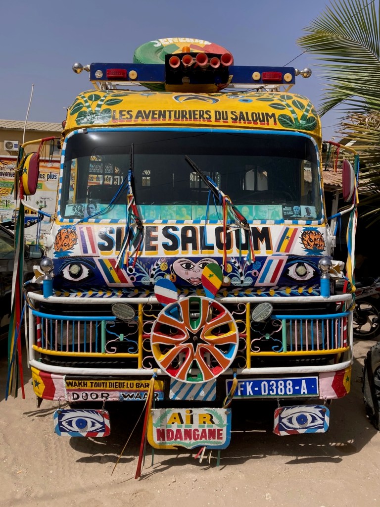 photo of the front of a small bus decorated in bright colors, streamers, flags, and signs saying "LES AVENTURIERS DU SALOUM" AND "SINE SALOUM"