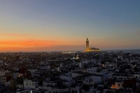 photo at sunset, looking across the rooftops of white-sided buildings with an illuminated mosque tower in the distance and orange clouds under an indigo sky