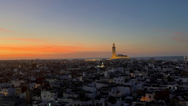 photo at sunset, looking across the rooftops of white-sided buildings with an illuminated mosque tower in the distance and orange clouds under an indigo sky