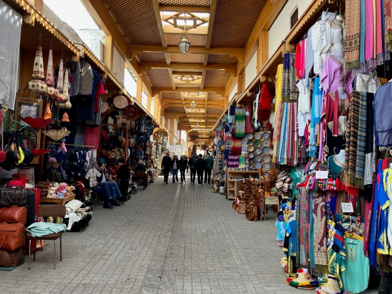photo of a narrow gray-brick street under an ornate wooden ceiling, lined on both sides with colorful clothing, rugs, ceramics, and other items for sale. Only 6 people are visible in the distance.
