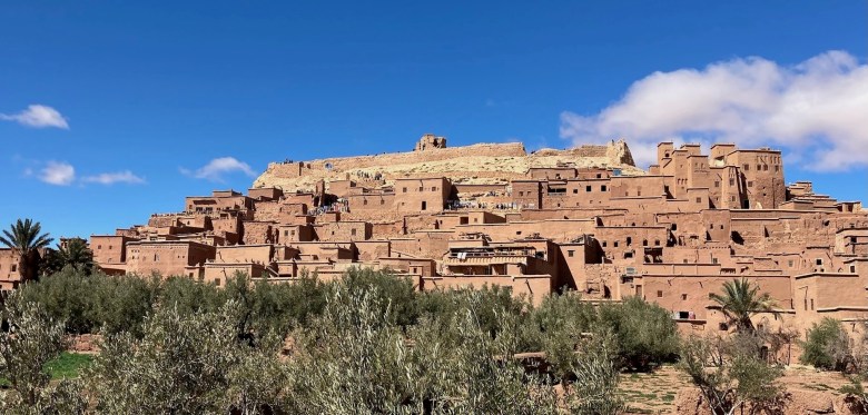 photo of sand-colored brick buildings, 1-3 stories high, clustered up the side of a hill, with green scrub gras in the foreground