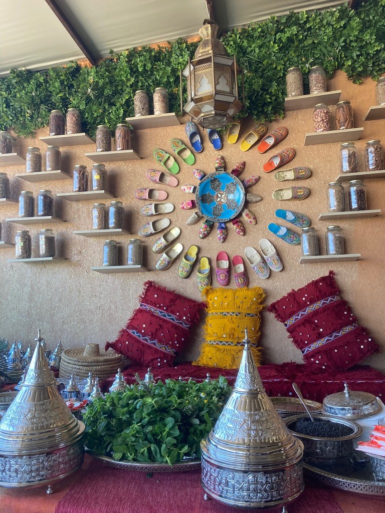 photo of a red table set holding silver serving dishes with cone-shaped lids, a big plate of greens, red and yellow pillows in the background, and a colorful array of slippers and spices on the wall