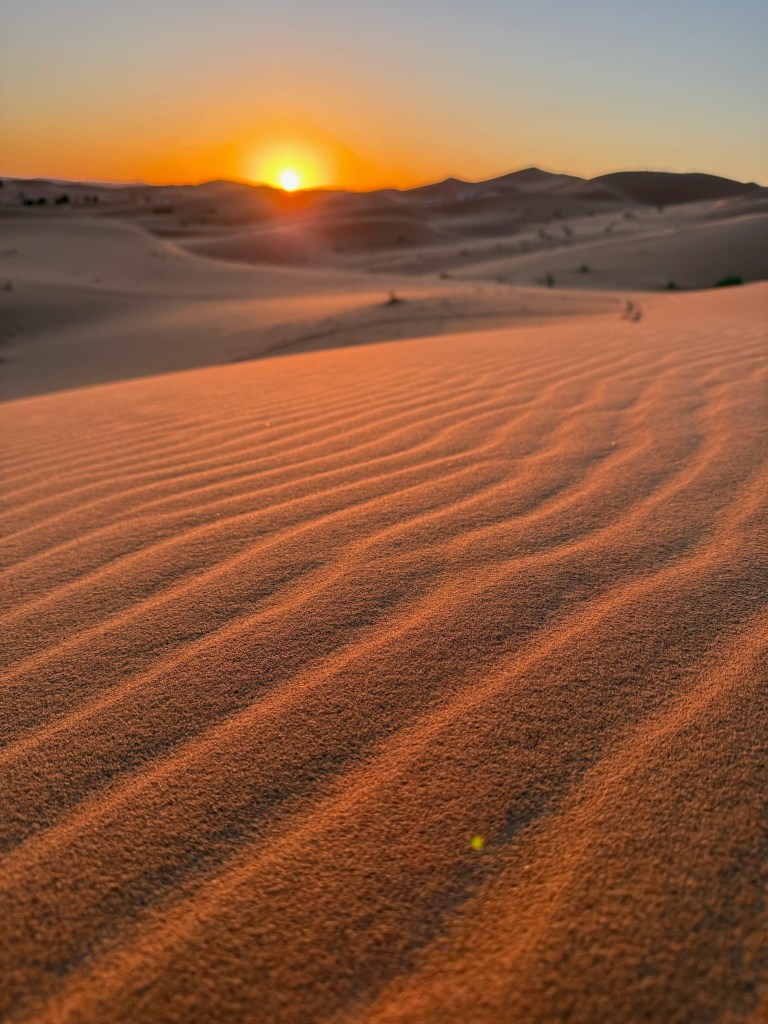photo of waves of brown sand in the foreground, dunes and an orange setting sun in the background